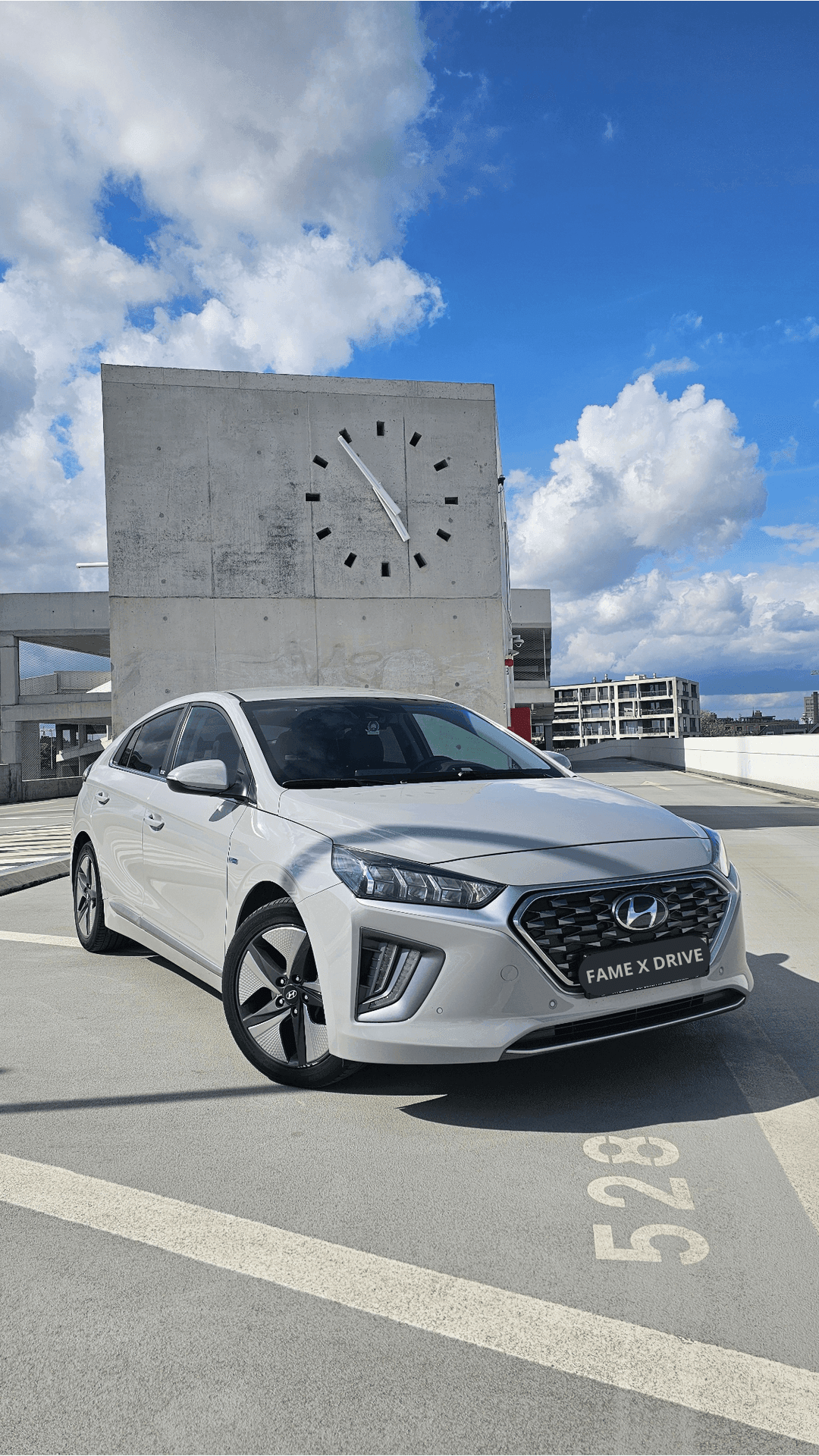 White Hyundai car parked on rooftop with large wall clock in the background under a partly cloudy sky.