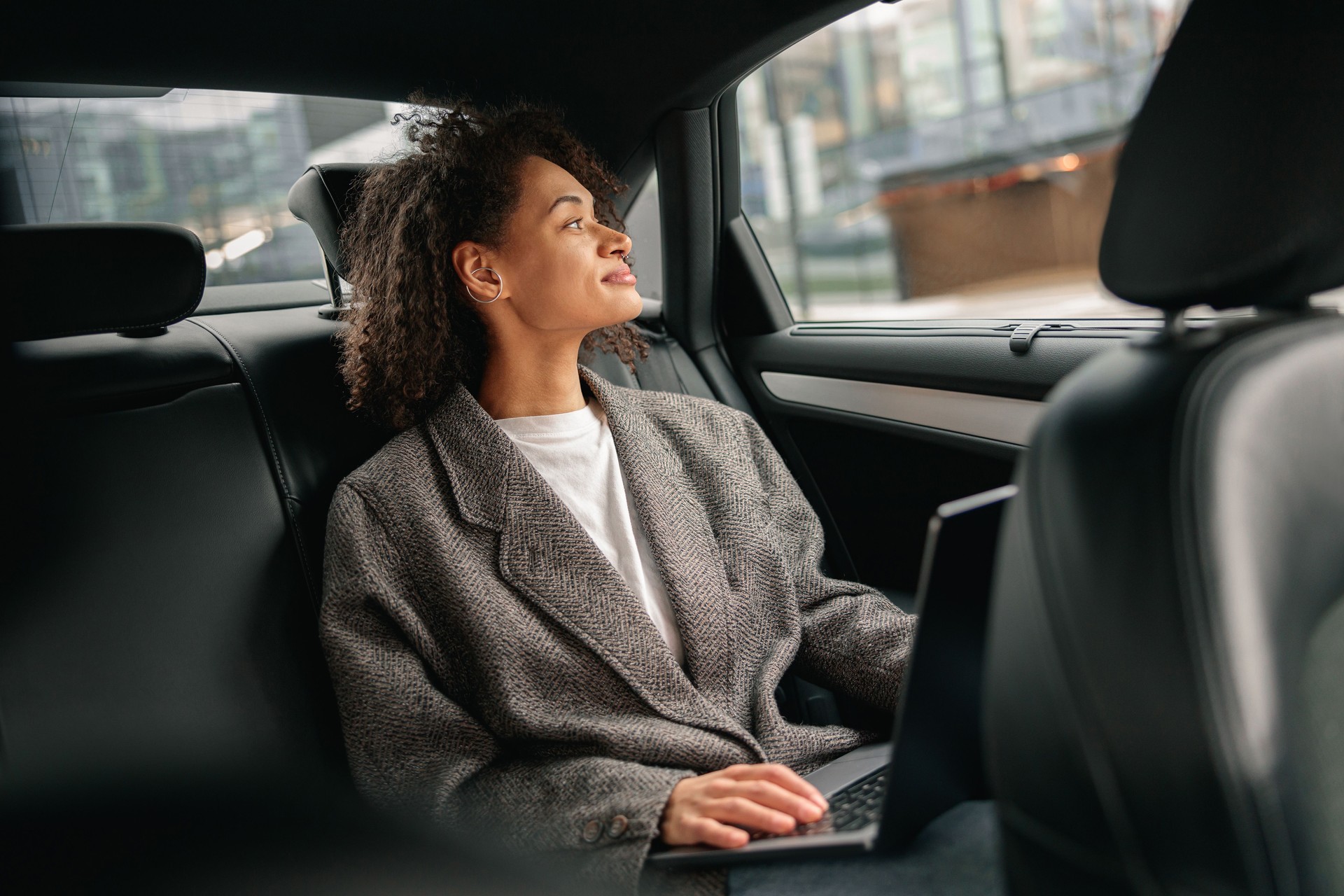 Woman freelancer working on laptop sitting on car backseat on way to office and looking at window